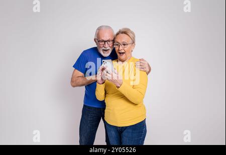 Aufgeregt schockiertes Seniorenpaar mit offenem Mund, das gute Nachrichten über Smartphones auf weißem Hintergrund liest Stockfoto
