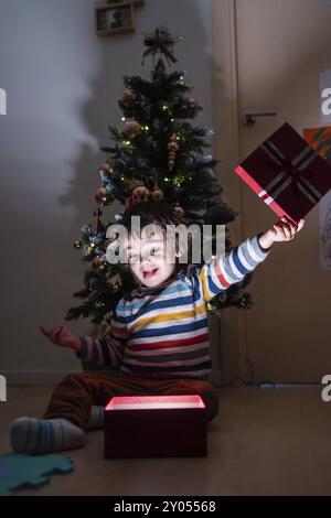 Ein kleiner Junge sitzt auf dem Boden vor einem Weihnachtsbaum und hält einen roten Kasten Stockfoto