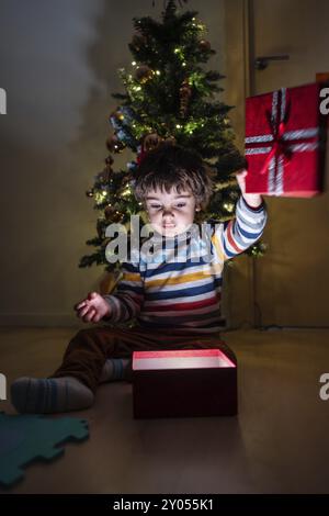 Ein kleiner Junge sitzt auf dem Boden vor einem Weihnachtsbaum und hält einen roten Kasten Stockfoto