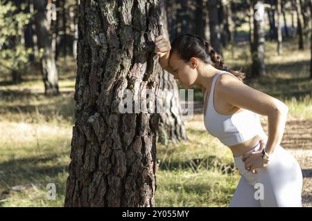 Ein junges, sportliches Mädchen, das nach dem Joggen oder Sport müde aussieht und sich an einen Baum lehnt Stockfoto