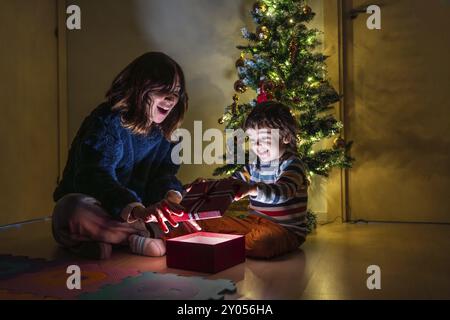 Ein Mädchen und ein Junge sitzen auf dem Boden vor einem Weihnachtsbaum. Das Mädchen öffnet ein Geschenk und der Junge beobachtet sie. Die Szene ist Joyfu Stockfoto