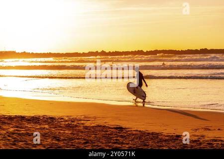 Ein Mann läuft am Strand mit einem Surfbrett. Der Himmel ist orange und das Wasser ist ruhig Stockfoto