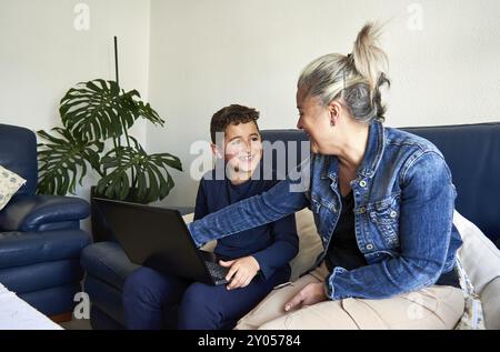 Eine Frau und ihr Sohn sitzen auf einem Sofa, lächeln und spielen mit einem Laptop in einem gemütlichen Wohnzimmer Stockfoto