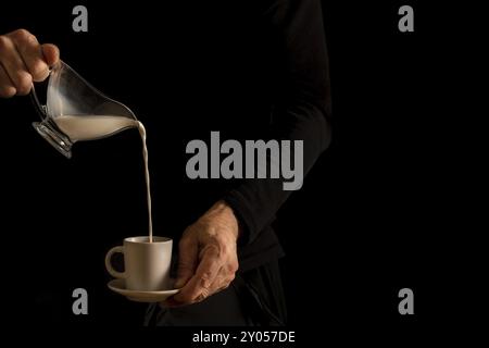 Die Hände eines nicht erkennbaren Mannes im Schatten, der Milch aus einer Glaskaraffe in eine Kaffeetasse gießt Stockfoto