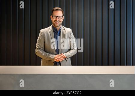 Porträt eines selbstbewussten männlichen Rezeptionisten mit Brille, der in die Kamera blickt und am Rezeptionstresen in der Lobby steht Stockfoto