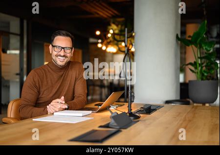 Porträt eines inspirierten männlichen Unternehmers in Brille, der über einem Laptop arbeitet, während er im Büro am Schreibtisch sitzt Stockfoto
