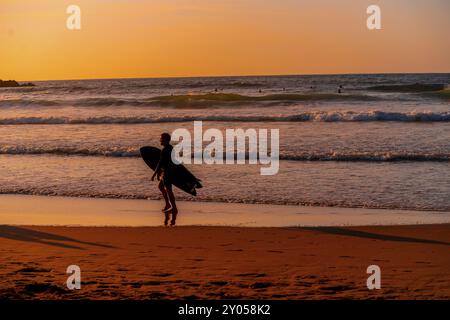 Ein Mann läuft am Strand mit einem Surfbrett. Der Himmel ist orange und das Wasser ist ruhig Stockfoto