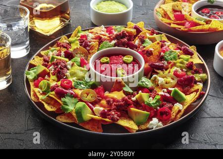 Nachos geladen. Mexikanische Nacho-Chips mit Rindfleisch, mit Guacamolensauce, Käsesalsa und Chili-Paprika, auf schwarzem Schiefergrund, mit Tequila-Getränken Stockfoto