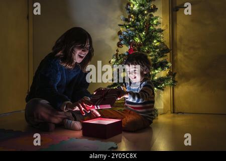 Eine Frau und ein Kind öffnen ein Geschenk vor einem Weihnachtsbaum. Die Szene ist fröhlich und festlich, da die beiden einen besonderen Moment miteinander teilen Stockfoto