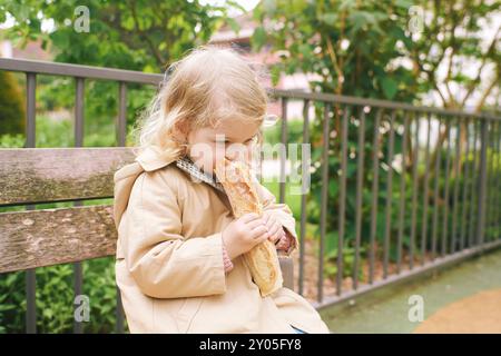 Süßes Porträt eines entzückenden kleinen Mädchens, das Baguette-Käsebrot isst und auf einer Bank im Park sitzt Stockfoto