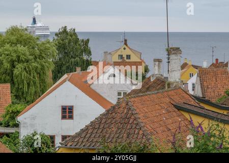 Meerblick über Häuser mit roten Dachziegeln, Schornsteinen, Bäumen und einem großen Schiff im Hintergrund, svaneke, bornholm, ostsee, dänemark, skandinavien Stockfoto