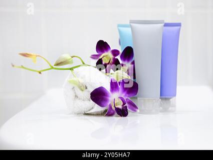 Spa-Set mit gewickeltem weißen Handtuch und wunderschönen lila Orchideenblüten neben bunten Kosmetikröhrchen auf weißem Tisch mit weißem Hintergrund Stockfoto