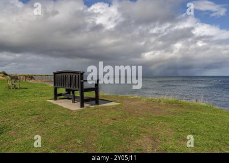 Eine Bank mit einem Regenbogen über der Nordsee Küste, im Benthall, Northumberland, England, UK gesehen Stockfoto