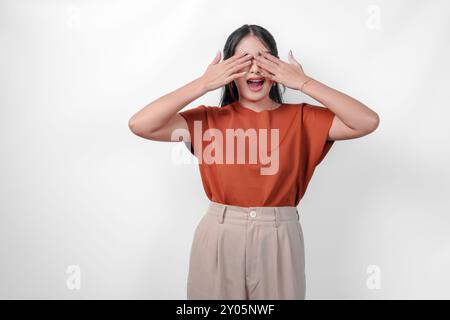 Eine junge asiatische Frau in einem braunen Hemd, die ihre Augen bedeckt und Angst hat, isoliert von weißem Hintergrund. Vorstellung von Überraschung oder Schock. Stockfoto