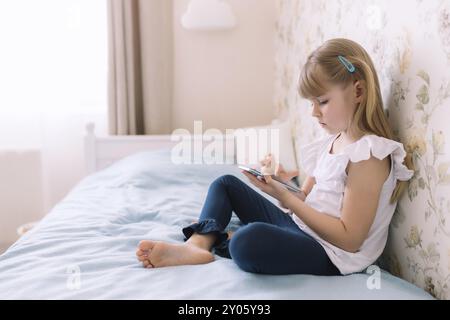 Ein kleines Mädchen sitzt auf dem Bett im stilvollen Schlafzimmer, hält das Telefon und liest etwas im Smartphone. Kommunikationskonzept Stockfoto