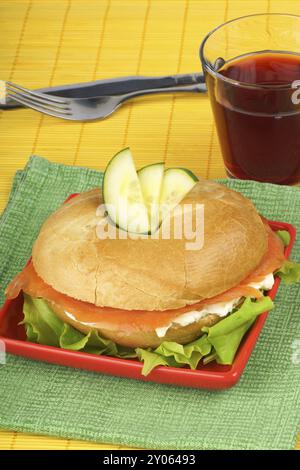 Bagel mit Weichkäse und Räucherlachs mit Gurkenscheiben serviert mit Salat auf einem roten Teller. Im Hintergrund ein Glas Preiselbeersaft, für Stockfoto