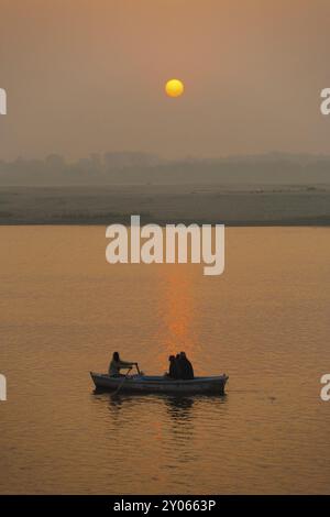 Ein Boot bringt Passagiere auf eine Bootstour bei Sonnenuntergang auf dem heiligen Ganges, während die Sonne von der Wasseroberfläche in Varanasi, Indien, Asien reflektiert wird Stockfoto