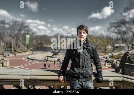 Ein junger, weißer, erwachsener Mann mit braunen Haaren steht vor einem Brunnen im Central Park New york, lehnt sich an einen Zaun, trägt eine Jacke und einen Schal und überwintert Stockfoto