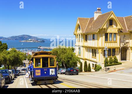 San Francisco, USA, 15. Mai 2016: Blaue Seilbahn mit hängenden Touristen, die auf der steilen Hyde St bergauf fahren, mit einem atemberaubenden Blick auf das Alcatraz Gefängnis, Bucht Wasser Stockfoto