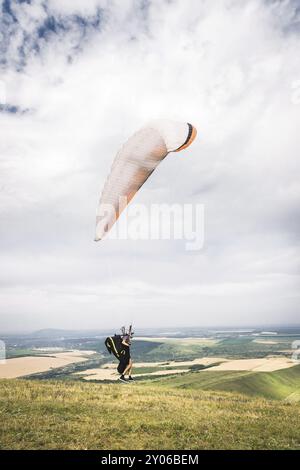 Ein Mann Gleitschirm, der vom Rand des Berges mit Feldern im Hintergrund abheben. Gleitschirmfliegen Stockfoto