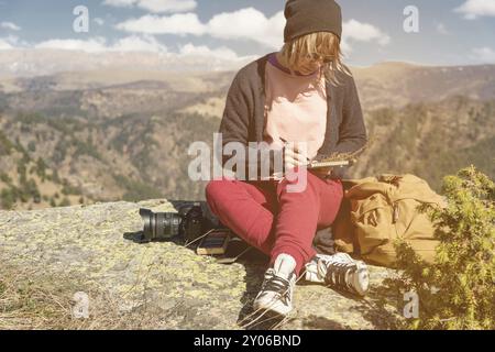 Porträt eines Hipster-Mädchens mit Sonnenbrille und Hut, das draußen auf einem Felsen in den Bergen vor blauem Himmel sitzt. Freiberuflicher Designer in Trav Stockfoto