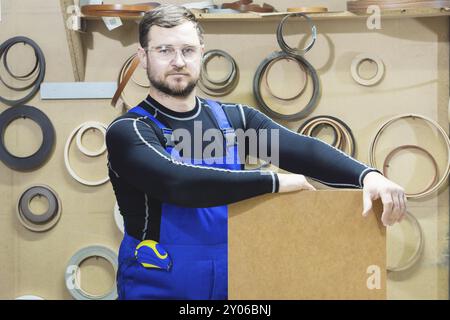 Der Hersteller von Möbeln in Spezialkleidung und Schutzbrille hält am Arbeitsplatz eine leere Platte für den Text. Handwerkliche Herstellung von Möbeln Stockfoto