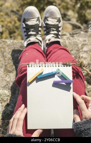 Blick von der ersten Person auf dem weiblichen Schoß ist ein Notizbuch mit Kreiden zum Zeichnen von Pastellfarben. Weibliche Hände, die ein Notizbuch in der Natur halten. Ein Mädchen sitzt Stockfoto