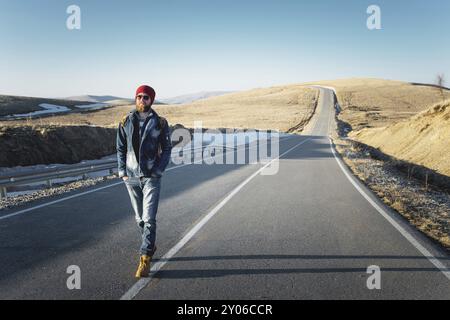 Ein stylischer bärtiger Hipster in Sonnenbrille mit einem Vintage-Rucksack spaziert an einem sonnigen Tag entlang der Asphaltstraße. Das Konzept des Anhalter- und Wanderns Stockfoto