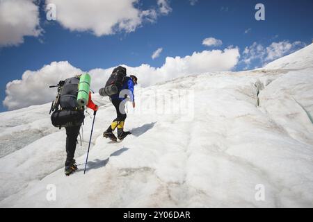 Zwei Touristen, ein Mann und eine Frau mit Rucksäcken und Steigeisen an den Füßen laufen entlang des Gletschers vor dem Hintergrund des Berges Elbrus und der Stockfoto