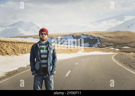 Ein stylischer bärtiger Hipster in Sonnenbrille mit einem Vintage-Rucksack steht an einem sonnigen Tag auf einem Landstraßenasphalt. Das Konzept des Anhalter- und Wanderns Stockfoto