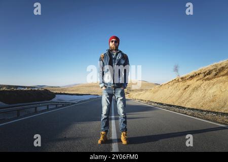 Ein stylischer bärtiger Hipster in Sonnenbrille mit einem Vintage-Rucksack steht an einem sonnigen Tag auf einem Landstraßenasphalt. Das Konzept des Anhalter- und Wanderns Stockfoto