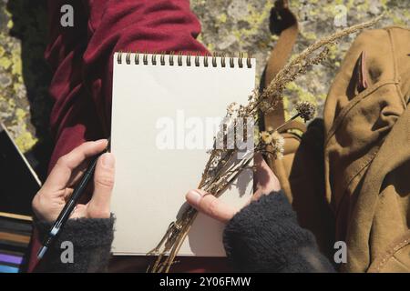 Nahaufnahme der Hände eines Mädchens, das ein leeres Notizbuch hält. Ein trockener Strauß Kräuter in der Hand und ein Bleistift. Reise-Designer-Künstler Stockfoto