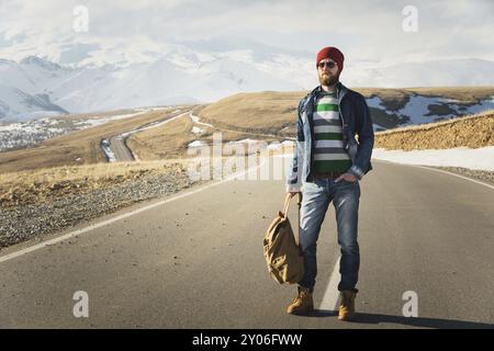 Ein stylischer bärtiger Hipster in Sonnenbrille mit einem Vintage-Rucksack steht an einem sonnigen Tag auf einem Landstraßenasphalt. Das Konzept des Anhalter- und Wanderns Stockfoto