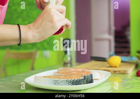 Frauenhände besprühen aus der Handmühle mit Gewürzen zwei rohe Steak vom Lachs, die auf einem weißen Teller in einer Küche in Grüntönen liegen. Das Konzept o Stockfoto