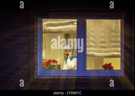 Eine Frau steht abends am hell beleuchteten Küchenfenster und spricht am Telefon Stockfoto