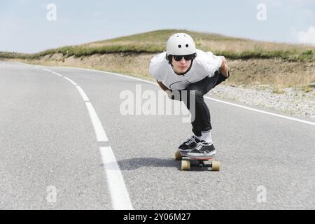 Der Typ mit Helm und Sonnenbrille fährt sehr schnell auf der Asphaltstraße in einem speziellen Sportständer sein Longboard Stockfoto