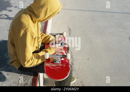 Nahaufnahme eines Teenagers, der in einem Sweatshirt mit Jeans und Turnschuhen sitzt und in einem Skatepark ein Telefon mit Kopfhörern und Skateboard hält Stockfoto