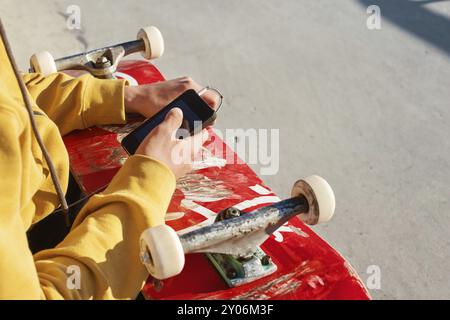 Nahaufnahme eines Teenagers, der in einem Sweatshirt mit Jeans und Turnschuhen sitzt und in einem Skatepark ein Telefon mit Kopfhörern und Skateboard hält Stockfoto