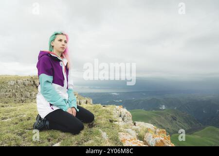 Ein Mädchen-Reisende mit bunten Haaren sitzt am Rande einer Klippe und blickt auf den Horizont auf einem Hintergrund eines felsigen Plateau Stockfoto