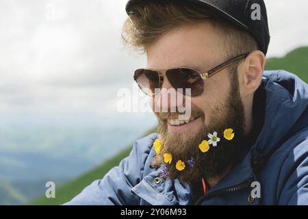 Nahporträt eines fröhlichen bärtigen Mannes in Sonnenbrille und grauer Mütze mit Wildblumen im Bart. Weiche Brutalität und gute Männlichkeit Stockfoto