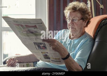 Eine alte Dame sitzt in ihrem Sessel am Fenster und liest die Tageszeitung Stockfoto