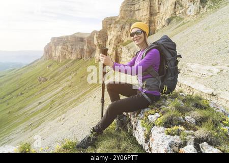 Lächelnde Reiseteilnehmerin mit gelbem Hut und Sonnenbrille, die am Fuße epischer Felsen mit einem Rucksack sitzt und in die Kamera schaut Stockfoto