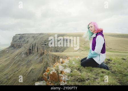 Ein Mädchen-Reisende mit bunten Haaren sitzt am Rande einer Klippe und blickt auf den Horizont auf einem Hintergrund eines felsigen Plateau Stockfoto