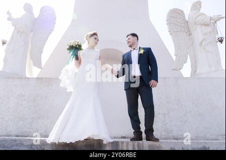 Porträt eines wunderbaren Paares, das an einem Hochzeitstag mit einem Blumenstrauß in der Hand vor dem Hintergrund eines orthodoxen christlichen Denkmals mit Engeln verbrachte. Stockfoto
