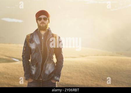 Modeporträt eines Lächelns bärtiger Hipster-jungen Mannes mit Sonnenbrille, Rucksack und Hut auf einem Hintergrund mit Copyspase in den Bergen bei Sonnenuntergang Stockfoto