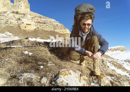 Porträt eines glücklichen Lachens Ein Hipster-Reisender mit Bart in Sonnenbrille sitzt und Schnürsenkel auf die Natur bindet. Ein Mann, der in den Bergen wandert Stockfoto