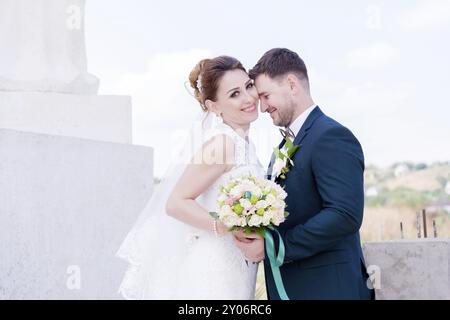 Porträt eines wunderbaren Paares, das an einem Hochzeitstag mit einem Blumenstrauß in der Hand vor dem Hintergrund eines orthodoxen christlichen Denkmals mit Engeln verbrachte. Stockfoto