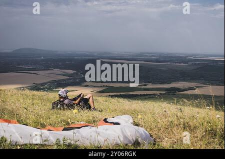 Ein professioneller Gleitschirm in voller Ausrüstung und ein Helm liegt und ruht auf dem Gras hoch in den Bergen und schaut auf die Wolken Stockfoto