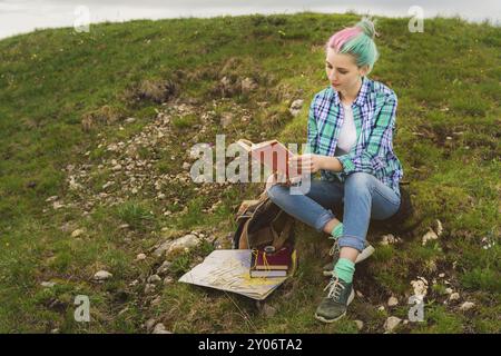 Ein Reisender sitzt in den Bergen auf dem Gras und liest ein Buch über den Hintergrund epischer Berge. Der Begriff des Lesens während der Ruhe und im Urlaub Stockfoto