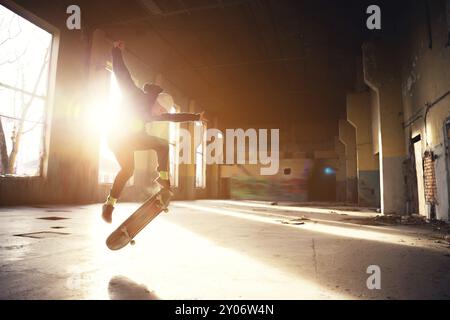 Ein junger Skater mit weißem Hut und schwarzem Sweatshirt macht einen Trick mit einem Skatesprung in einem verlassenen Gebäude im Hintergrund der untergehenden Sonne. Die Stockfoto